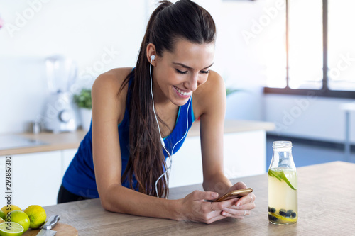 Sporty young woman listening to music with mobile phone after training in the kitchen at home.