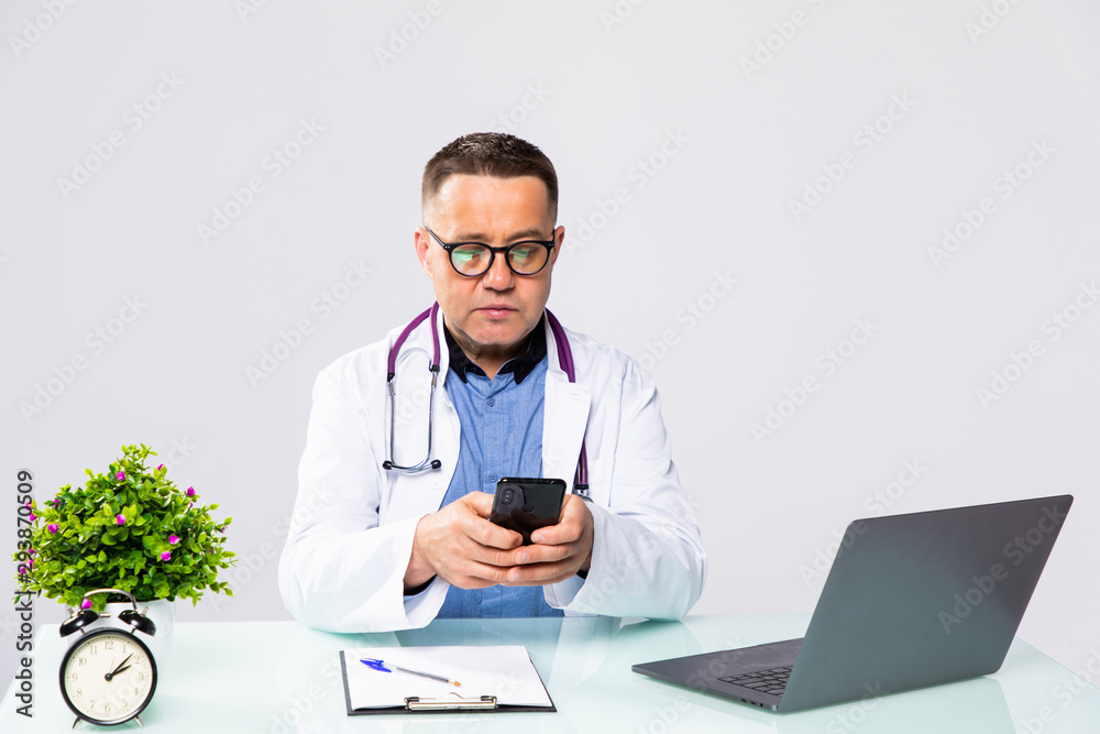 Smiling male doctor using mobile smart phone and working on laptop computer in medical room in the hospital, electronic health records system EHRs, teleconference or telemedicine concept. Smiling male doctor using mobile smart phone and working on laptop computer in medical room in the hospital, electronic health records system EHRs, teleconference or telemedicine concept.