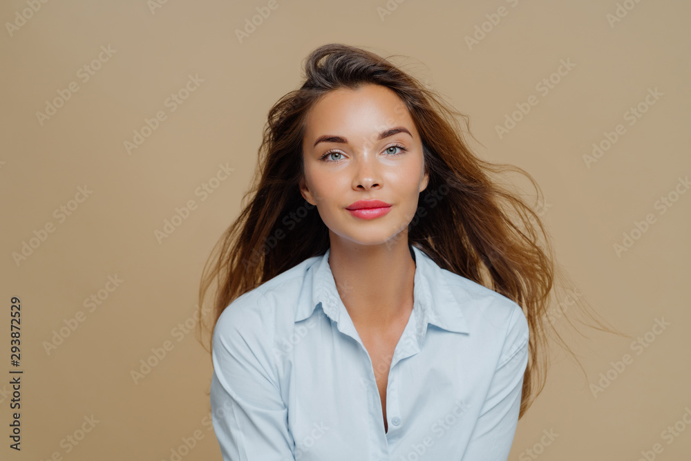 Lovely young female has long wavy hair floating in wind, dressed in elegant blue shirt, wears lipstick and makeup, poses against brown background, has direct gaze at camera, healthy facial skin Lovely young female has long wavy hair floating in wind, dressed in elegant blue shirt, wears lipstick and makeup, poses against brown background, has direct gaze at camera, healthy facial skin