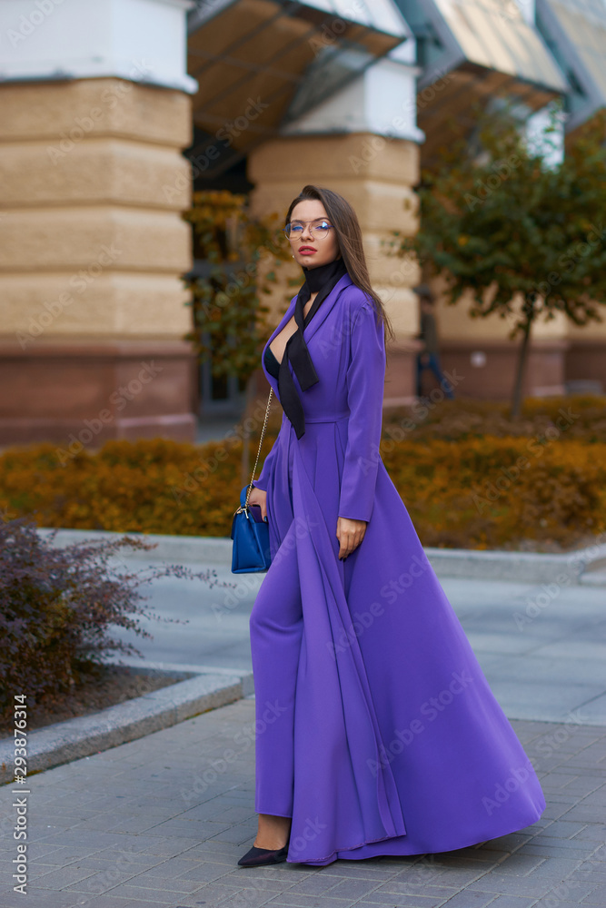 Young beautiful caucasian woman with long straight hair and makeup in purple trousers and long coat cape standing and posing at city street in autumn