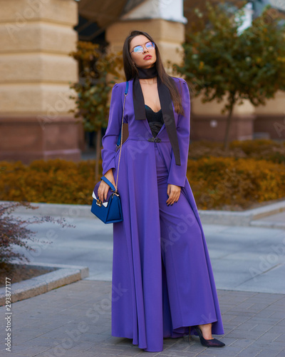 Young beautiful caucasian woman with long straight hair and makeup in purple trousers and long coat cape standing and posing at city street in autumn © Dmitry Tsvetkov