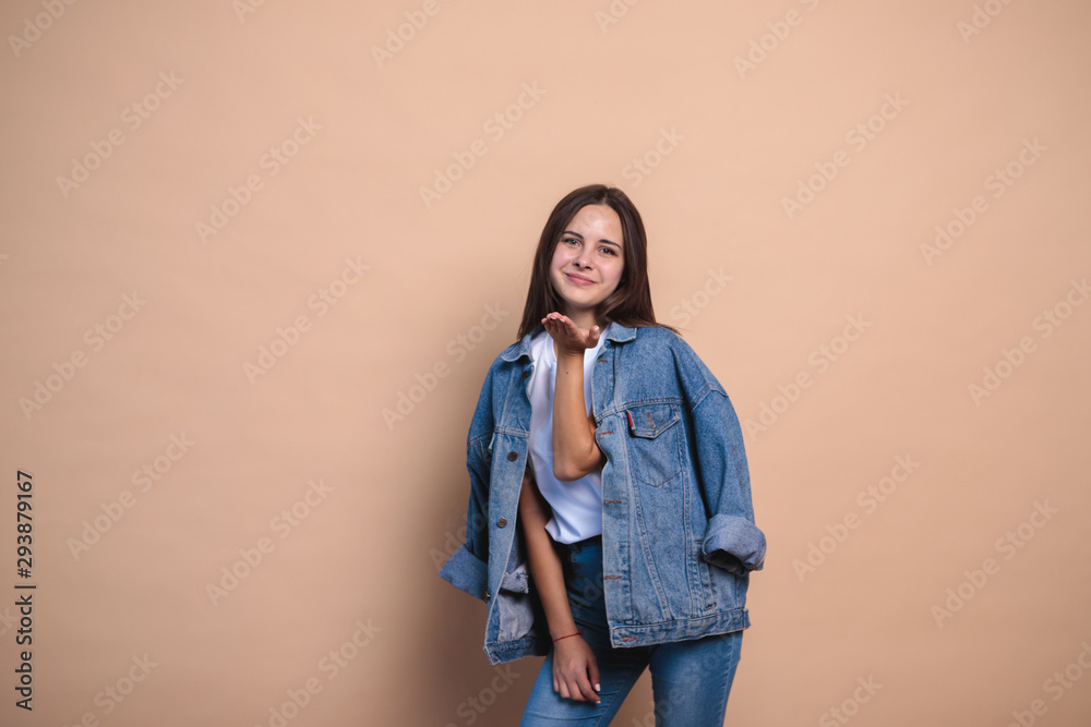 stylish girl in jeans clothes on a light background, stands and looks at the camera and blows a kiss. space for text