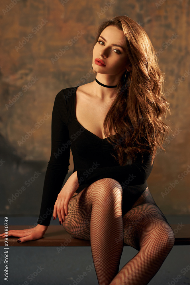 Half length portrait of young sexy brunette woman with wavy hair in black  body and fishnet tights sitting on bemch in grunge gym. Fashion 90's style  shoot Stock Photo | Adobe Stock