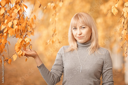 a woman with blond hair in a gray jacket and jeans, shot against the autumn foliage on a cloudy day