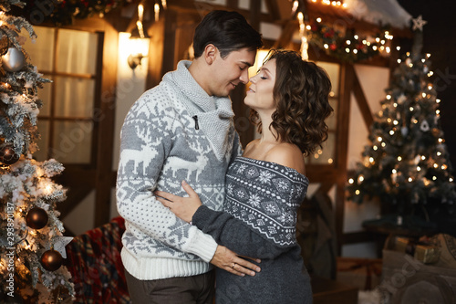Beautiful couple of lovers in Scandinavian sweaters at New year interior. Sexy model girl and handsome man looking to each other and posing near Christmas tree