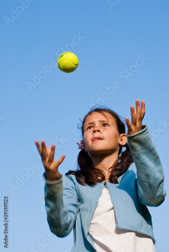 Cute young girl throwing and catching a tennis ball against a blue sky