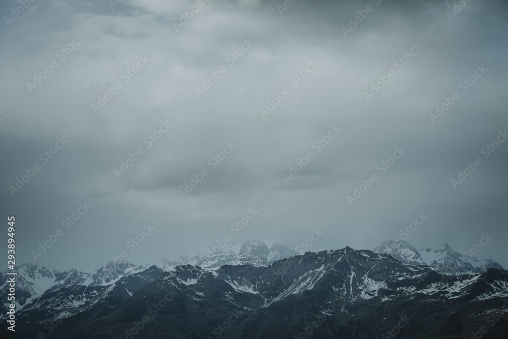 Storm is coming with dark grey clouds over the horizon of the snowy peaks of the Italian Alps in bottom part of an image and bigger part of negative space in upper part