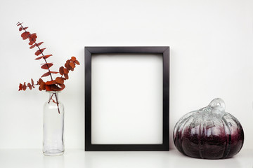 Mock up black frame with fall red eucalyptus branch and pumpkin decor on a shelf or desk. Autumn concept. Portrait frame against a white wall.