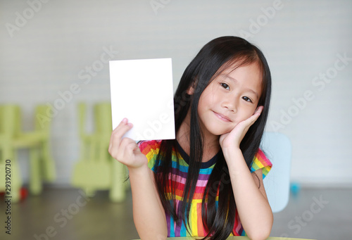 Happy little Asian child girl holding blank white paper card in her hand. Kid showing empty paper note copy space in children room.