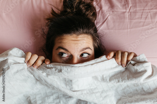 Scared and surprised young woman with open eyes hiding face under blanket, pretty frightened and curious girl feeling shy peeking from duvet, covering with white sheet, head shot close up. Top view