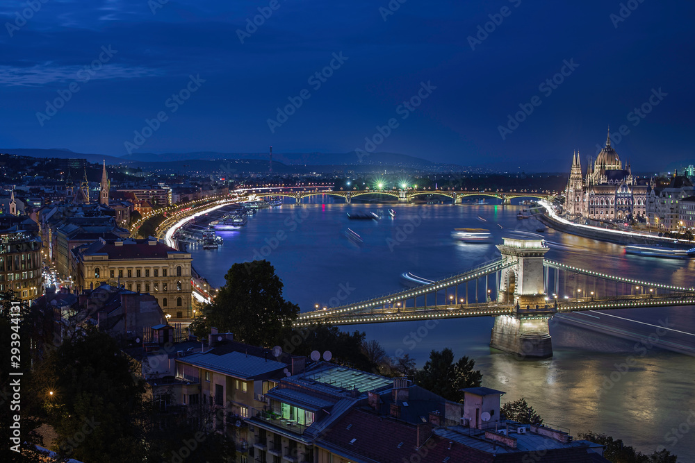 Budapest, Hungary - Illuminated Szechenyi Chain Bridge on a night photograph with Parliament of Hungary, moving ships on River Danube and clear dark blue sky after sunset