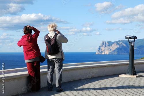 Elderly couple take pictures of beautiful sea and mountains landscape, back view. Active old age, traveling, enjoying life.