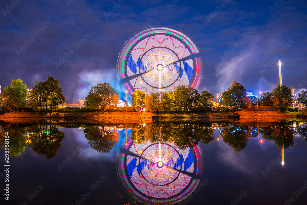 Germany, Illuminated colorful big wheel in motion and rollercoaster of popular folk festival called cannstatter wasen in stuttgart bad canstatt by night reflecting in water