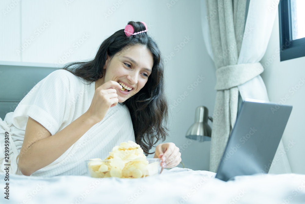 A beautiful woman working and listening to music on a laptop with yellow headphone and lying down on the bed at a condominium in the morning.