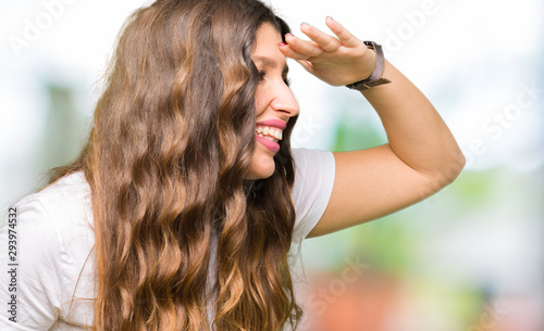 Young beautiful woman wearing casual white t-shirt very happy and smiling looking far away with hand over head. Searching concept.