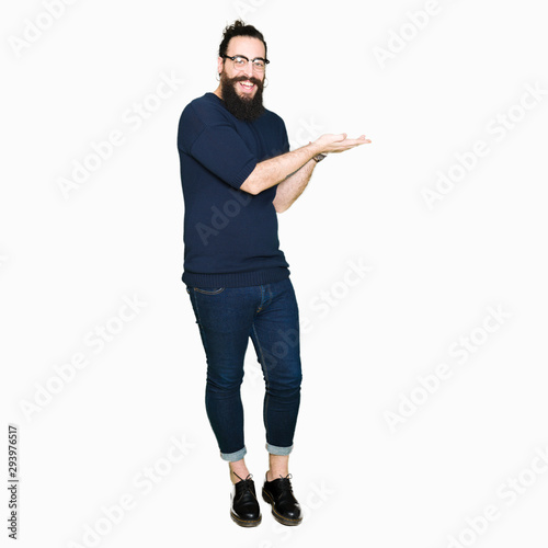 Young hipster man with long hair and beard wearing glasses Pointing to the side with hand and open palm, presenting ad smiling happy and confident