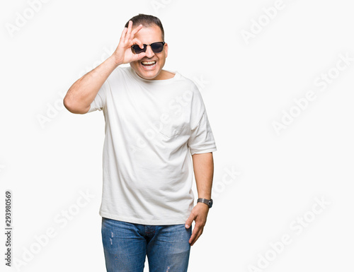 Middle age arab man wearig white t-shirt and sunglasses over isolated background doing ok gesture with hand smiling, eye looking through fingers with happy face.