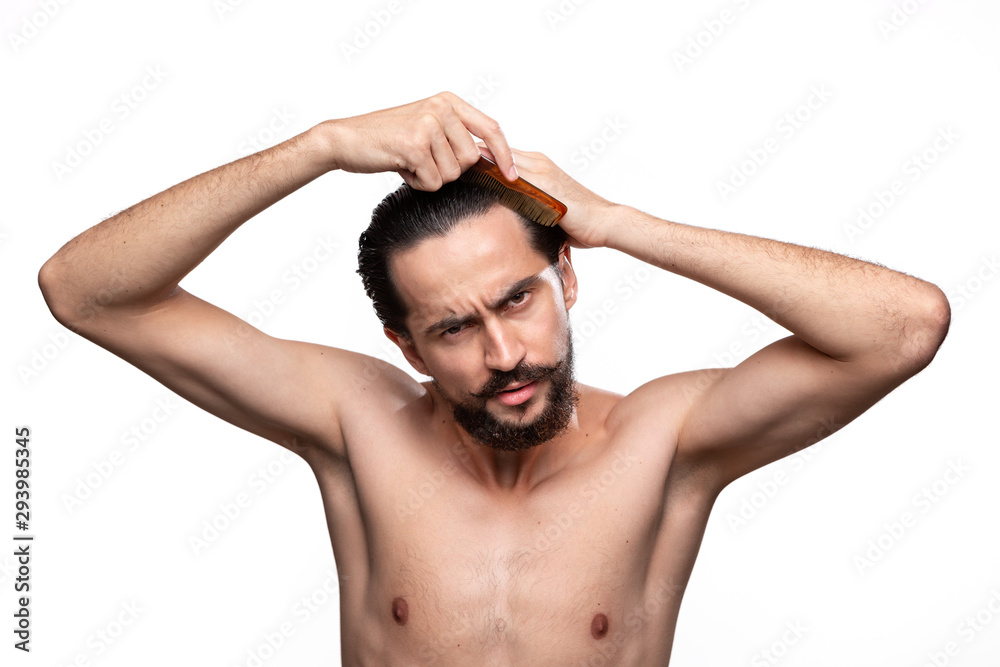 Stylish handsome bearded with mustaches combing hair standning bare isolated over white background. Concept of morning treatment. Morning routine