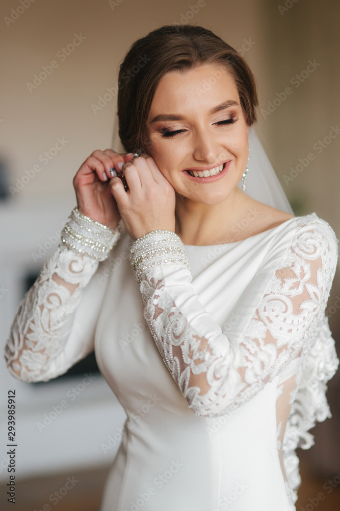 Close up portrait of beautiful bride standing by the window at home and tying on earrings. Charming bride in white wedding dress Close up portrait of beautiful bride standing by the window at home and tying on earrings. Charming bride in white wedding dress