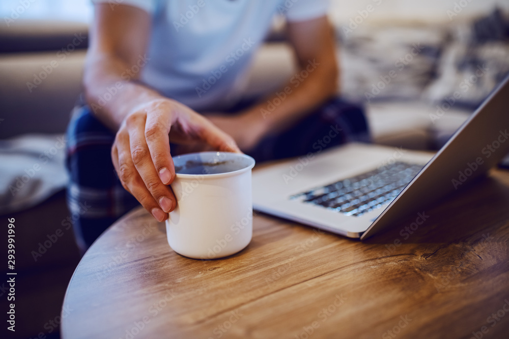 Close up of man taking mug with morning coffee from desk. Man is dressed in pajamas, sitting on sofa in living room and using laptop. Weekend activities. Selective focus on hand. Close up of man taking mug with morning coffee from desk. Man is dressed in pajamas, sitting on sofa in living room and using laptop. Weekend activities. Selective focus on hand.