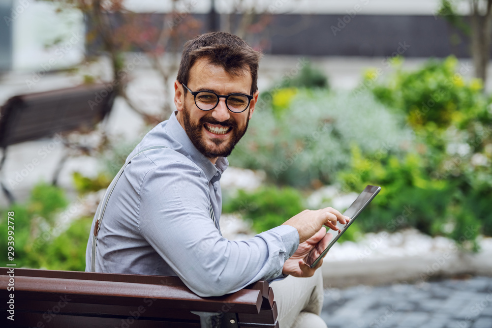 Obraz premium Smiling attractive caucasian businessman with eyeglasses sitting on bench in park and using tablet while looking over shoulder.