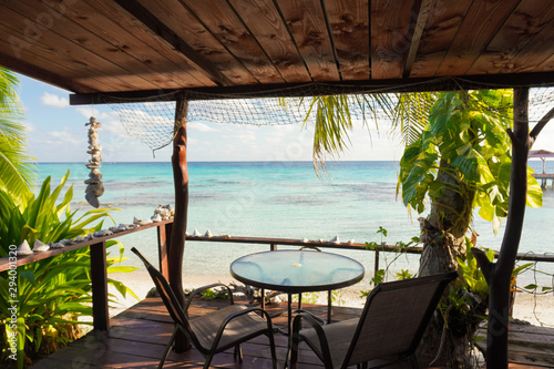 Fototapeta Naklejka Na Ścianę i Meble -  View of a blue tropical lagoon from the wooden deck of a bungalow with a table and chairs and framed by palm trees and seashells