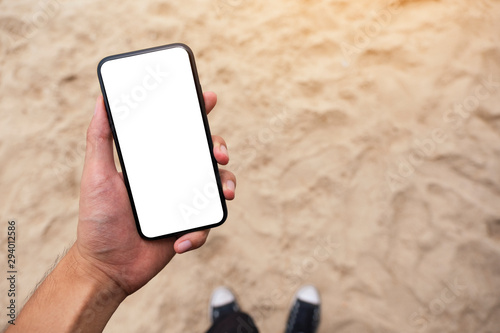 Mockup image of a man's hand holding black mobile phone with blank desktop screen while standing on the beach