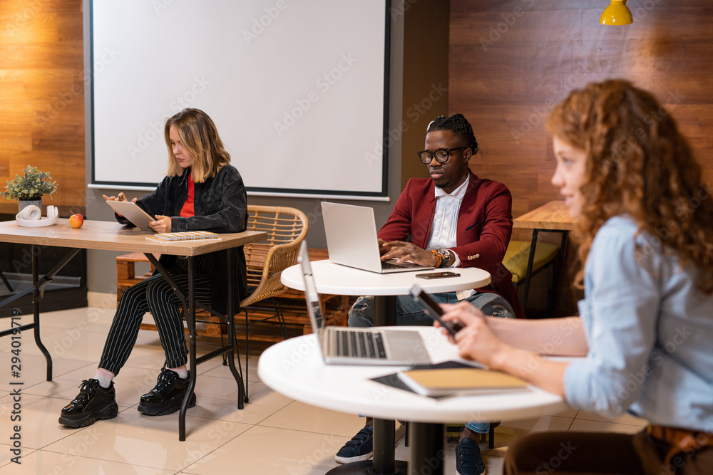 Group of young intercultural students of college sitting by tables in cafe