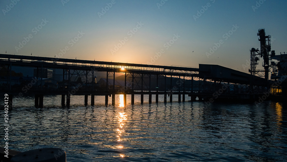 Obraz premium Sunrise at sunrise over the seaport with a calm sea surface on a cloudless autumn morning against the background of buildings for loading grain.