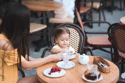 Mother with baby daughter sitting in cafe eating tasty dessert and drinking tea outdoors. Motherhood. Summer season. Childhood. Breakfast time.