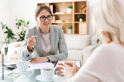 Confident young auditor with pen sitting in front of client and consulting her photo
