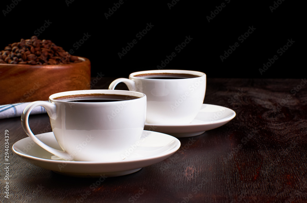 Two cups of coffee in saucers on a dark wooden table. In the background, a wooden dish with coffee beans on a tea towel. Two cups of coffee in saucers on a dark wooden table. In the background, a wooden dish with coffee beans on a tea towel.