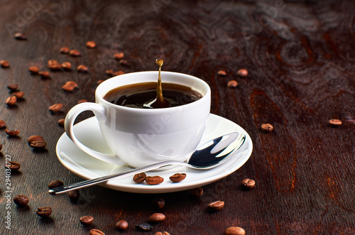 A Cup of coffee on a saucer and coffee beans on a dark wooden table. A drop of coffee splashed in the Cup.