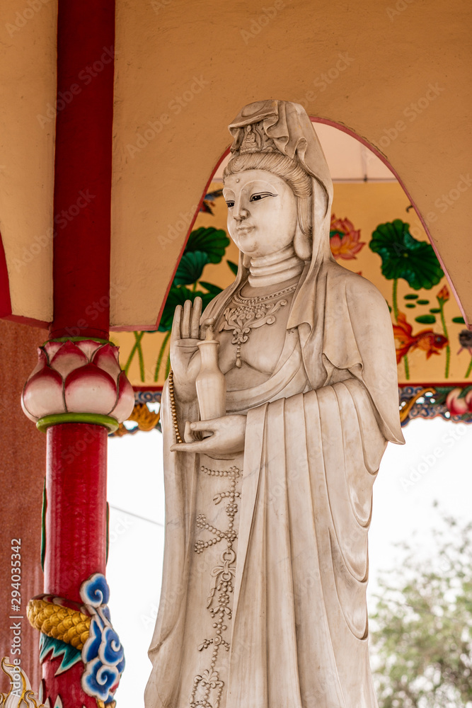 Fototapeta premium Si Racha, Thailand - March 16, 2019: Closeup of Gray statue of Guan Yin at her open circular shrine on Ko Loi Island. Backed by reds, beiges and whites.