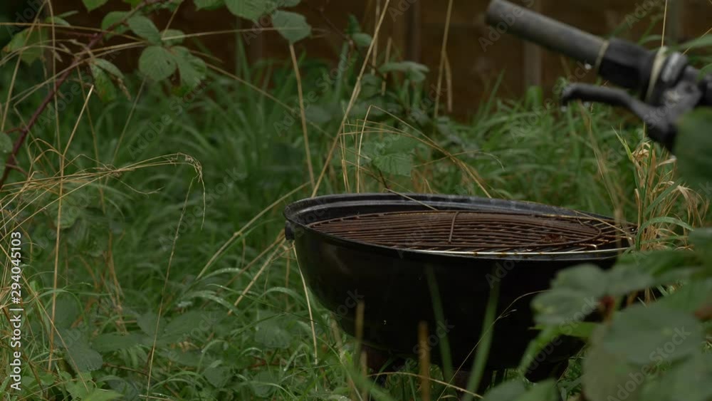 Overgrown garden with rusty BBQ and bike handle in the rain