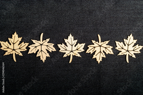 Line with five delicate light brown wooden leaves on dark grey textile material background, top view with space for text on the right side, flat lay with laser cut wooden objects