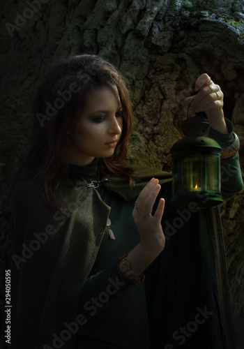 Young witch with green gloak and candle stay in forest