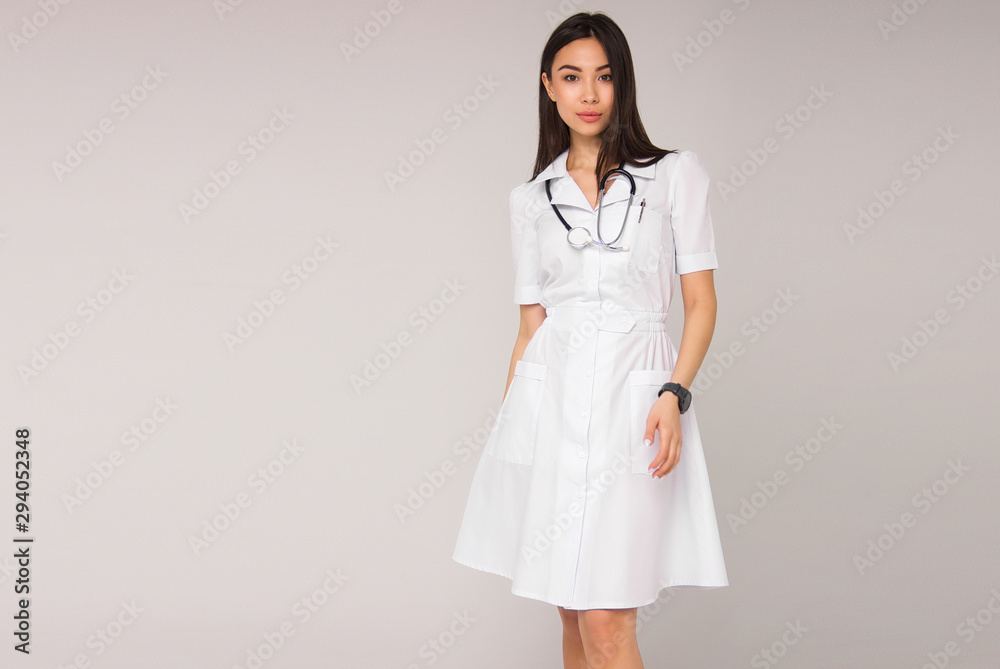 young stylish brunette nurse in white medical gress with stethoscope on her neck is standing on white wall background and smiling cute. medical fashion concept. free space on left side young stylish brunette nurse in white medical gress with stethoscope on her neck is standing on white wall background and smiling cute. medical fashion concept. free space on left side