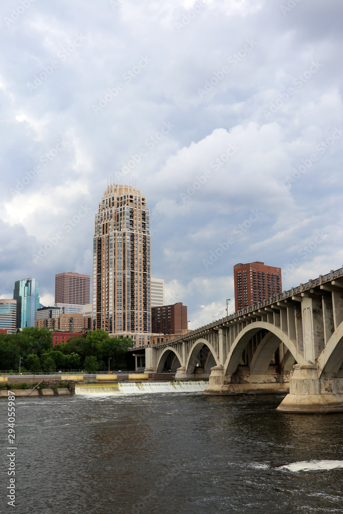 Naklejka premium Urban cityscape and modern architecture background. Cloudy sky over Minneapolis downtown skyline and Third Avenue Bridge above Mississippi river, vertical composition. Midwest USA, Minnesota.