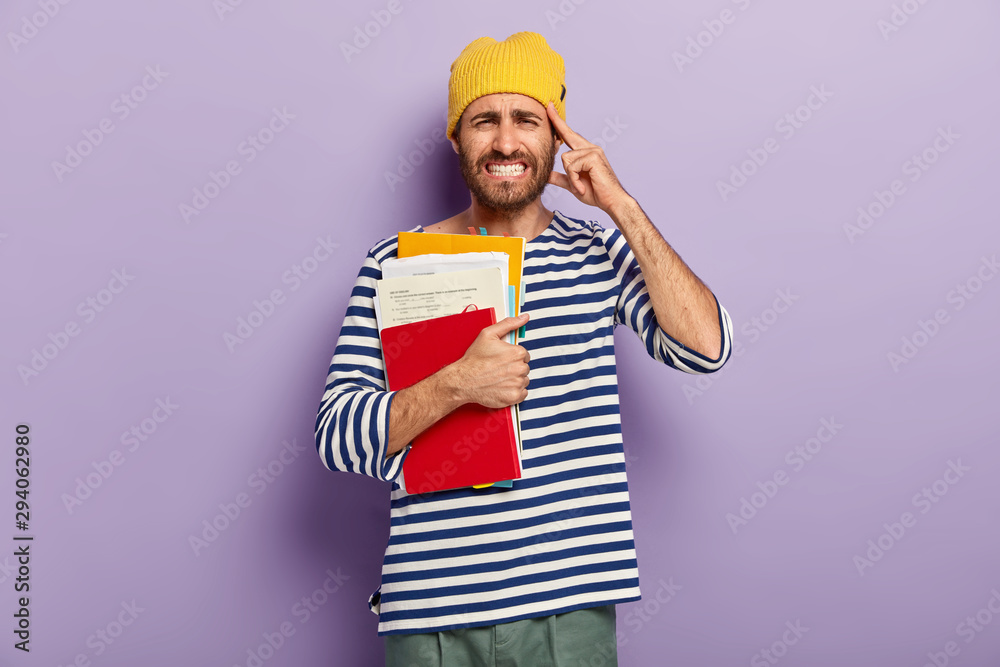 Photo of displeased male student clenches teeth, feels pain in temple, holds papers and textbooks, has upset facial expression, dressed in casual striped jumper, poses against purple studio wall