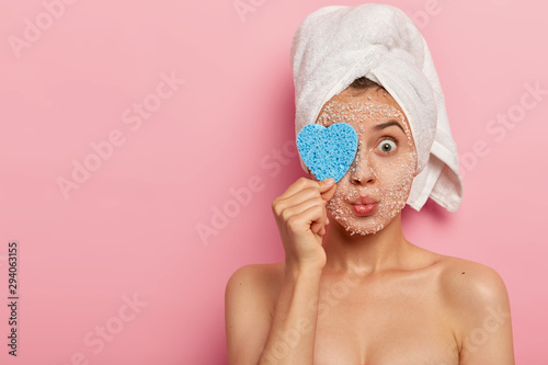Isolated shot of pleasant looking woman covers eye with sponge, keeps lips rounded, has naked well cared body, bugged eyes, wears white towel, isolated over pink background, blank space aside