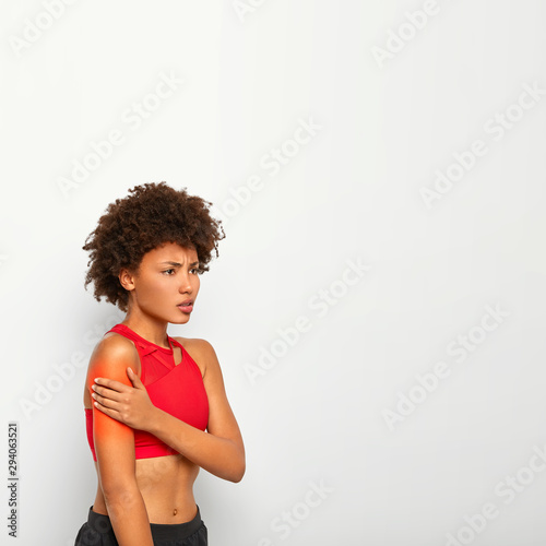 Vertical shot of displeased African American woman suffers from pain in shoulder, has bare belly, wears red top, poses against white background, holds hand on arthritic place, isolated on white