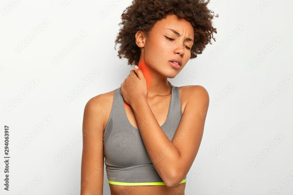 Cropped image of upset curly haired young woman suffers from neck pain, being injured during sport training, wears casual grey top, focused down, isolated over white background. Health problems Cropped image of upset curly haired young woman suffers from neck pain, being injured during sport training, wears casual grey top, focused down, isolated over white background. Health problems