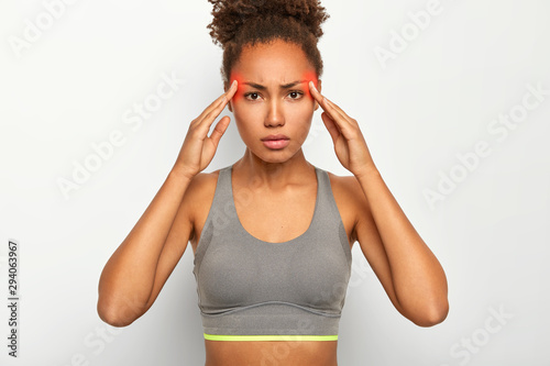 Image of serious black woman touches both temples, suffers from migraine, wears grey top, has red marked zones on head showing painful area, looks tensed and tired, isolated over white background