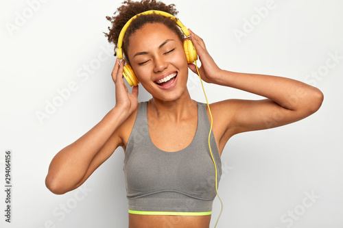 Emotional beautiful Afro American woman with curly hair, laughs positively, enjoys loud music in headphones, keeps eyes closed from pleasure, wears grey top, goes in for sport with favourite track