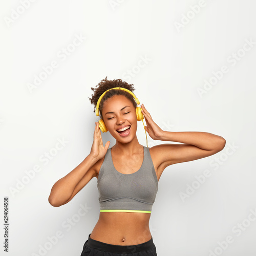 Vertical shot of positive dark skinned woman enjoys loud music in stereo headphones, sings songs pleasantly, keeps eyes closed, has fitness training, poses over white background, blank space above