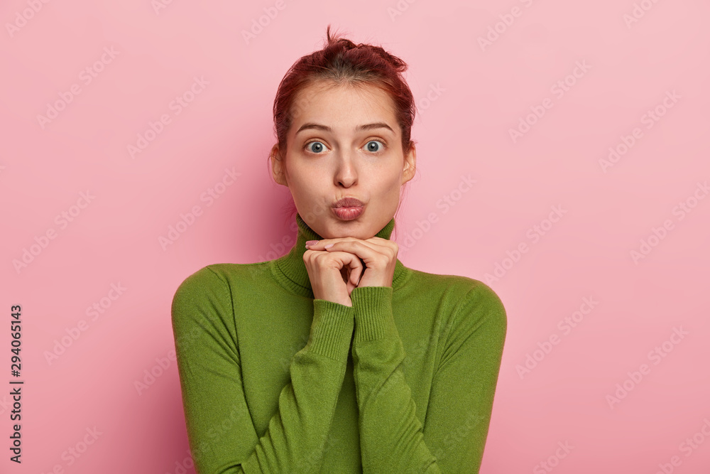 Photo of lovely European woman pouts lips, wants to kiss someone, wears green  turtleneck, has no makeup, keeps both hands under chin, makes grimace at  camera, isolated over pink studio wall. Stock