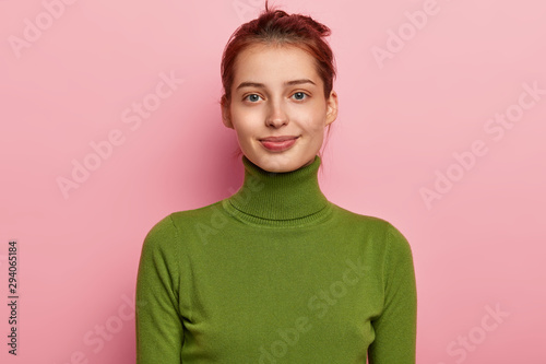 Portrait of beautiful young lady has dark combed hair, appealing appearance, wears casual green turtleneck, looks gladfully into camera, poses against pink background, feels pleased to have day off