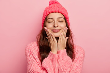 Portrait of pleased European woman touches both cheeks with palms, wears knitted hat and sweater, closes eyes with satisfaction, imagines future date with boyfriend enjoys winter time isolated on pink