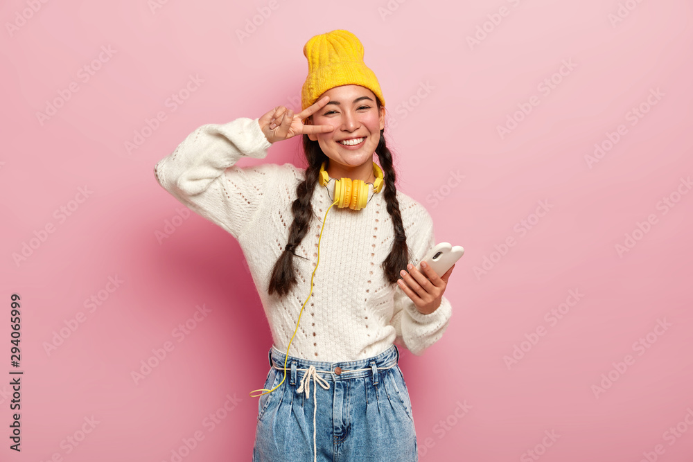 Smiling young korean woman makes peace gesture over eye, holds modern cell phone, has two plaits, smiles gently, wears yellow hat and jeans, poses against pink background. People and technology Smiling young korean woman makes peace gesture over eye, holds modern cell phone, has two plaits, smiles gently, wears yellow hat and jeans, poses against pink background. People and technology